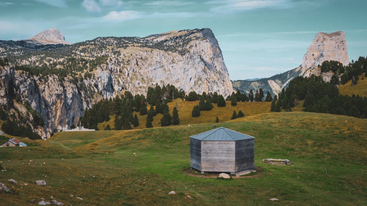 A wooden Mountain hut in the middle of vast green meadows, surrounded by majestic mountains.