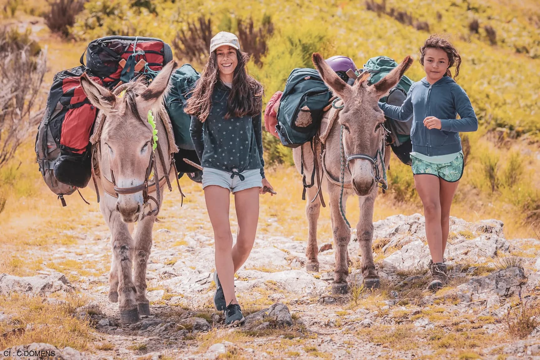 Twee wandelaars en hun beladen ezels, wandelend over een zonnig pad in de Cevennen.