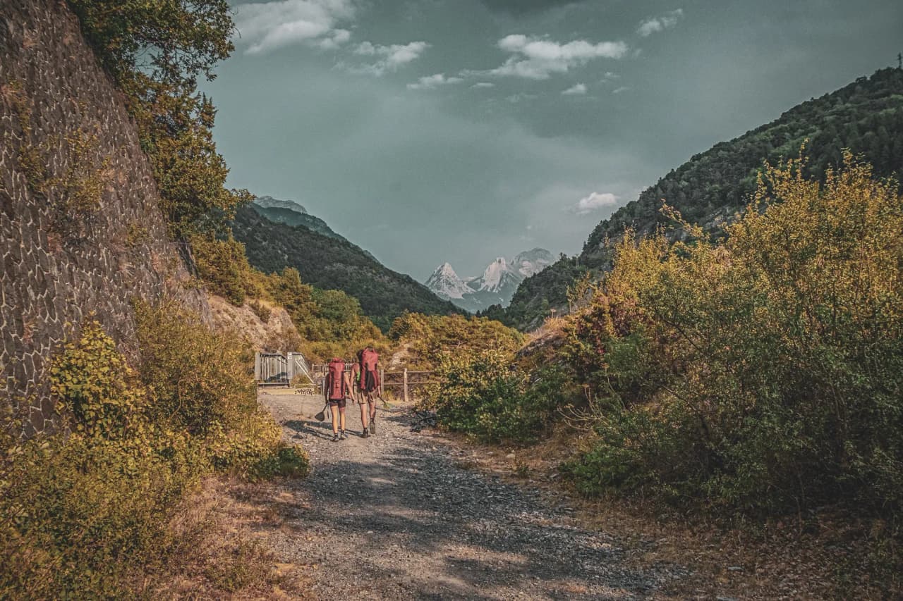 Two hikers follow an alpine path, surrounded by majestic mountains and greenery.