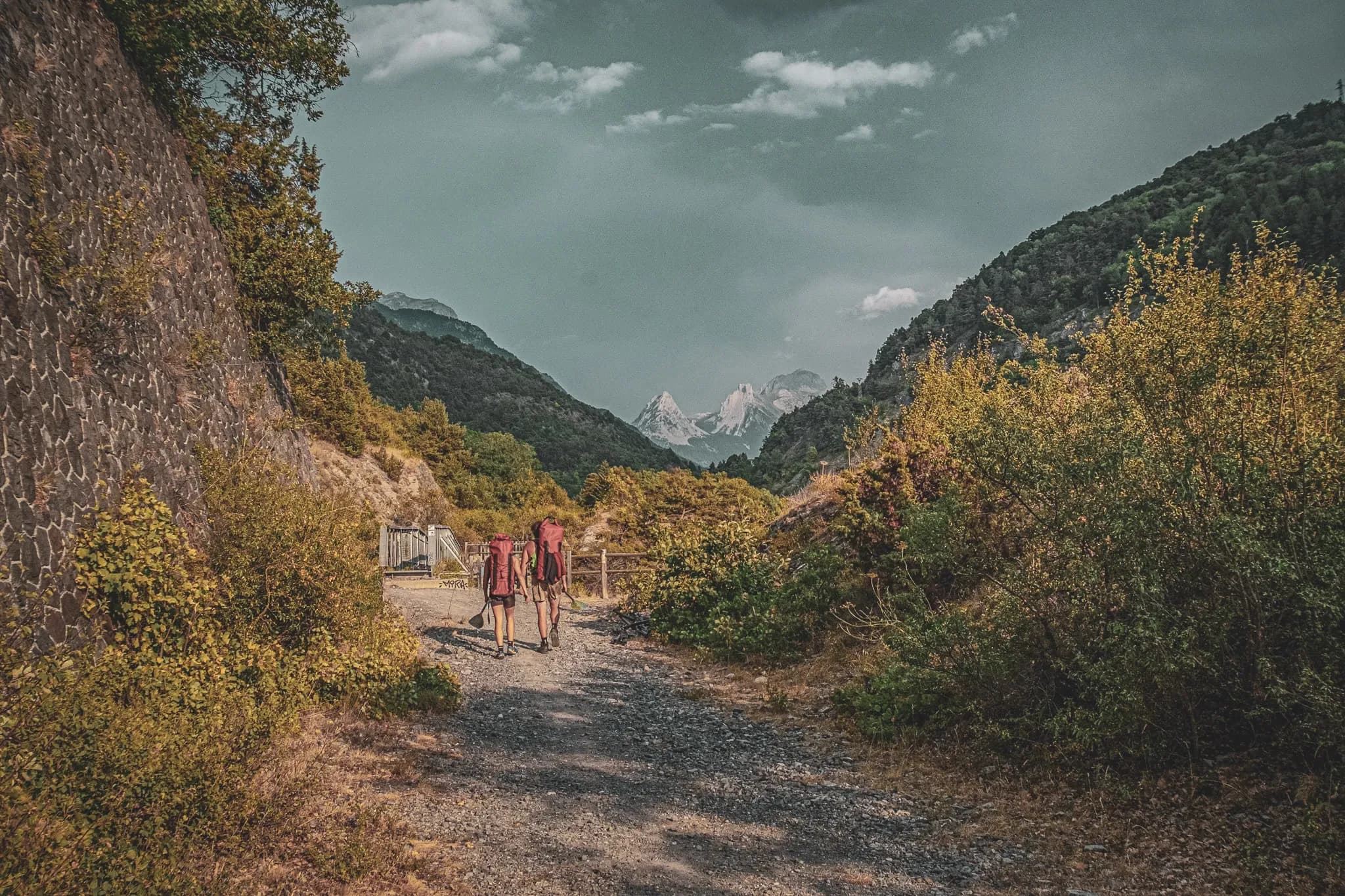 Two hikers follow an alpine path, surrounded by majestic mountains and greenery.