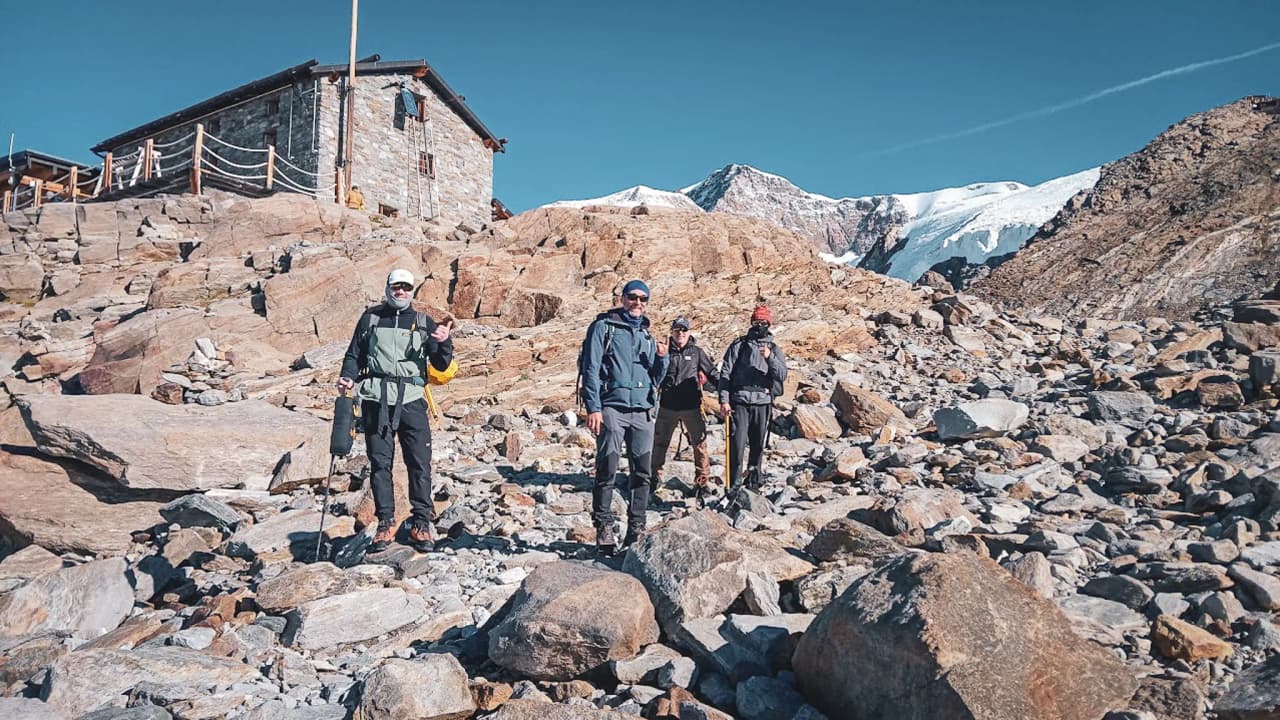 Four adventurers are posing on rocky ground, with the Margherita mountain hut and the snow-capped peaks in the background.