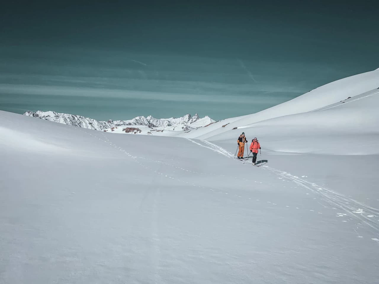 Twee skiërs trekken door een majestueus besneeuwd landschap, met de bergen op de achtergrond.