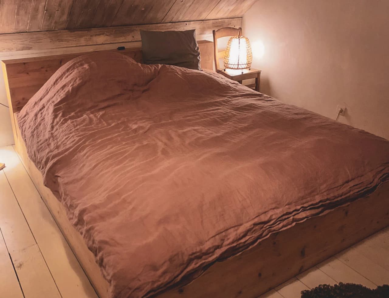Een ruim bed met een bruinachtig dekbed staat in een kamer met een schuin dak. Het bed is gemaakt van hout, met een simplistisch frame dat het natuurlijke materiaal benadrukt. Daarnaast staat een lamp met een rustiek ontwerp