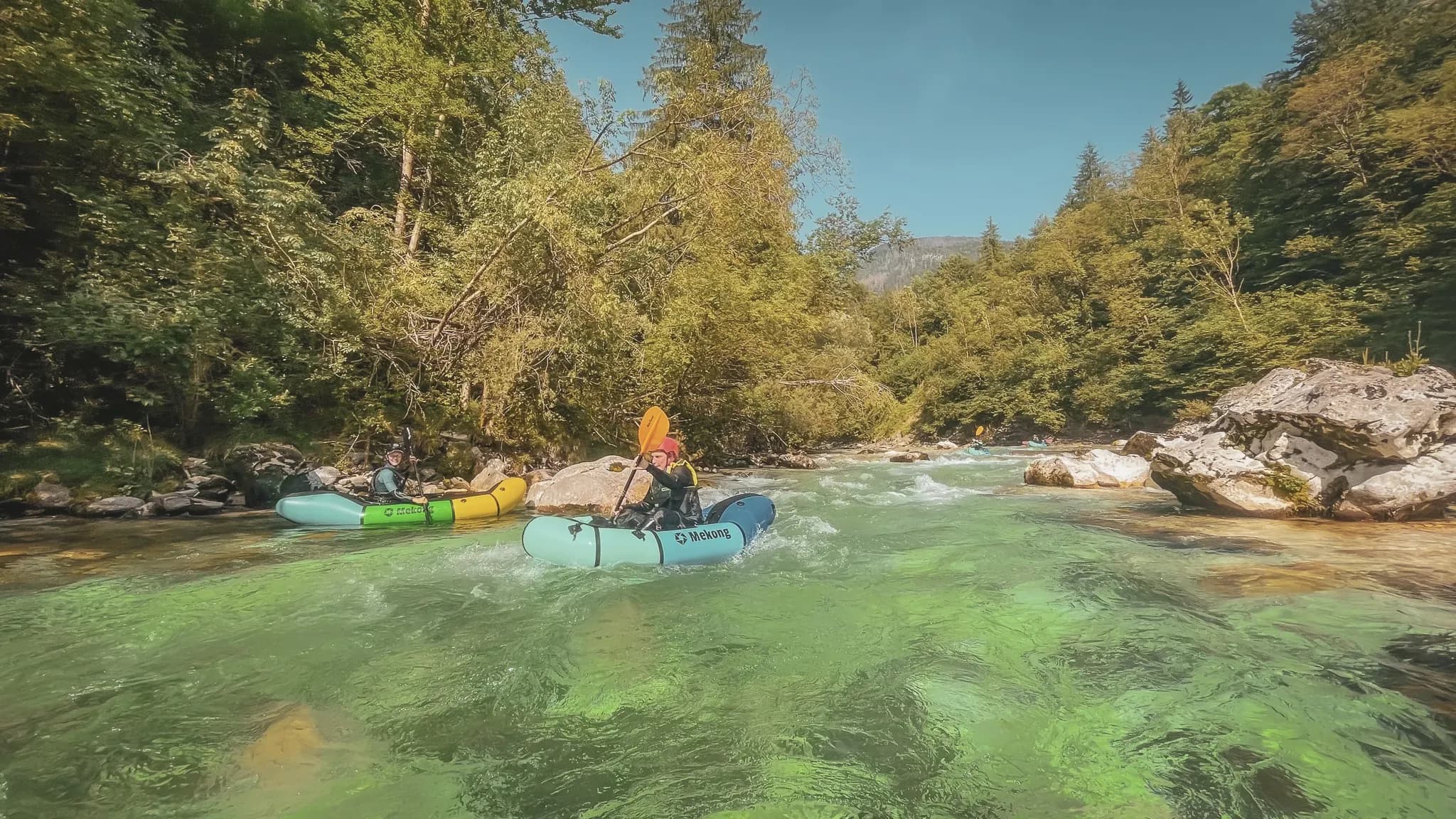 Two packraft adventurers navigate a turquoise river surrounded by lush greenery.