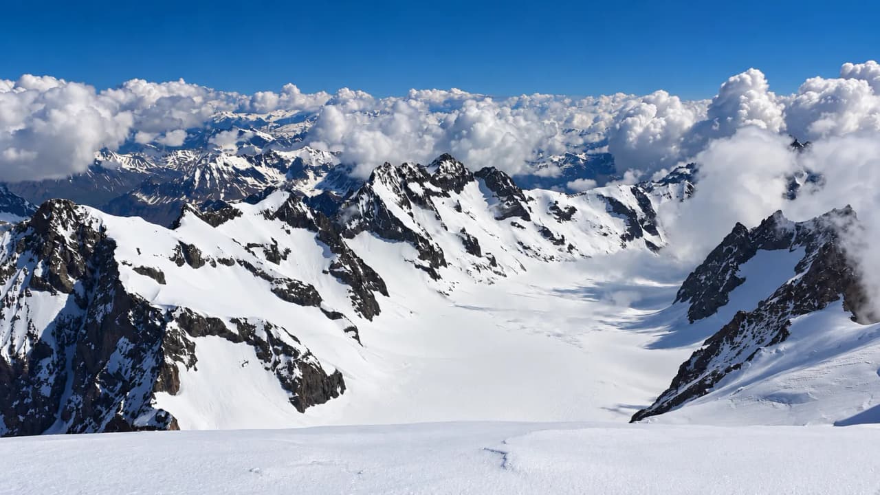 Uitzicht vanaf een besneeuwde top over een uitgestrekte gletsjer en een wolkenzee boven het Écrins-massief.
