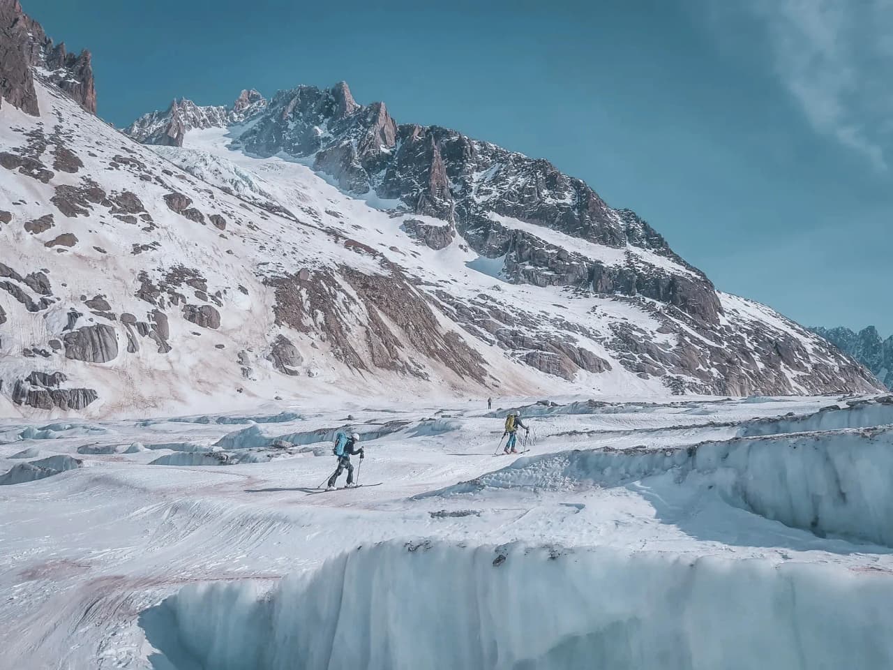 Skieurs traversant un glacier majestueux sous un ciel bleu, entourés de montagnes grandioses.