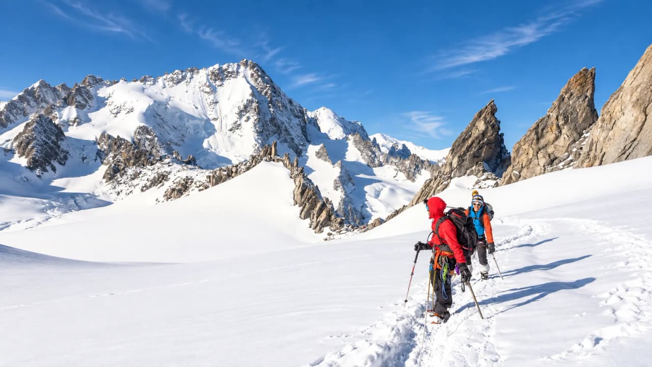 Twee bergbeklimmers in touwgroep op een besneeuwde gletsjer, tussen granieten pieken onder een blauwe lucht.
