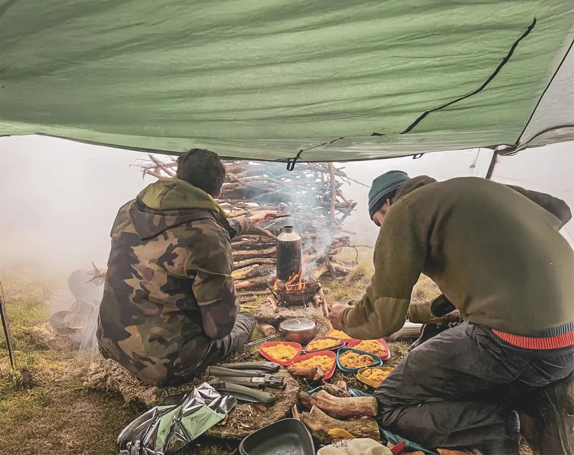 Two adventurers cook under a tarpaulin, surrounded by nature, in the heart of the Vercors forests.