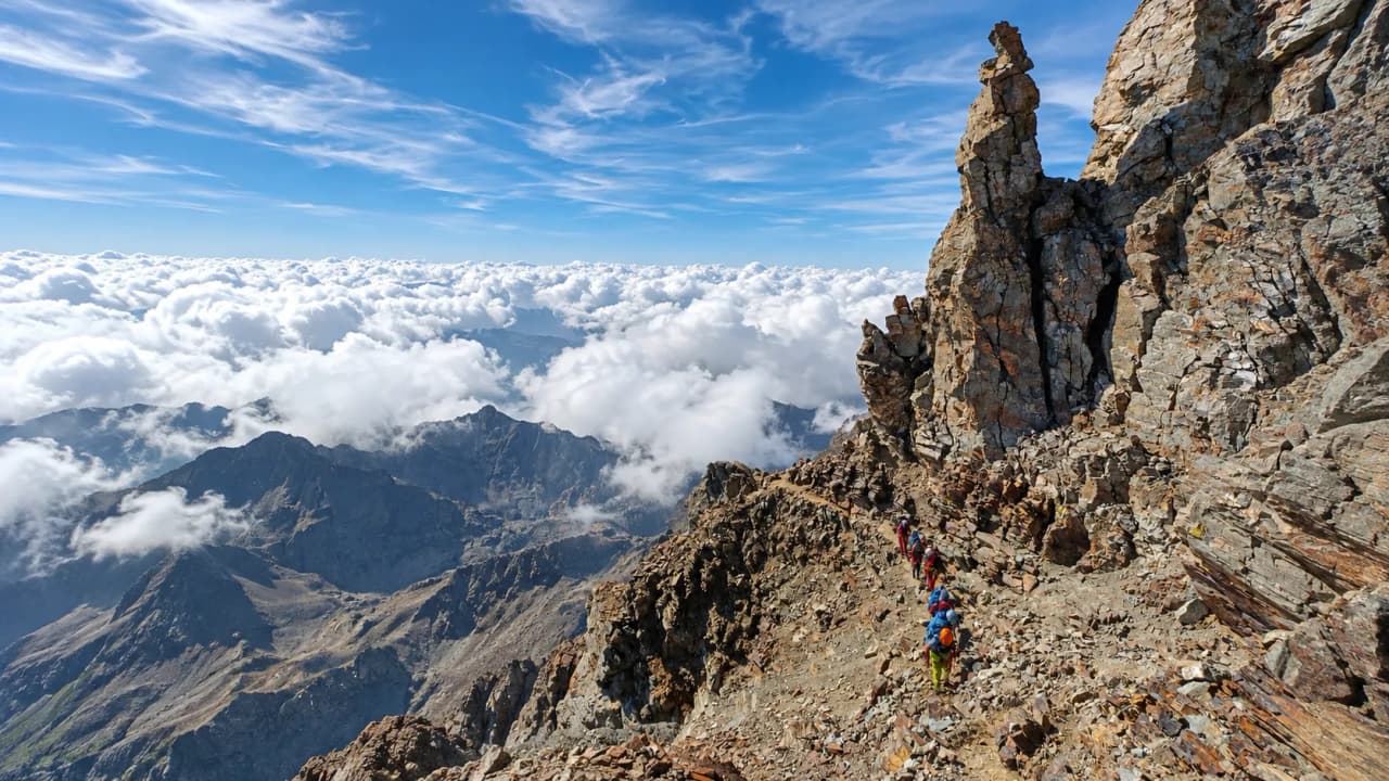 A group of walkers on an alpine trail, with a majestic landscape of mountains and clouds.