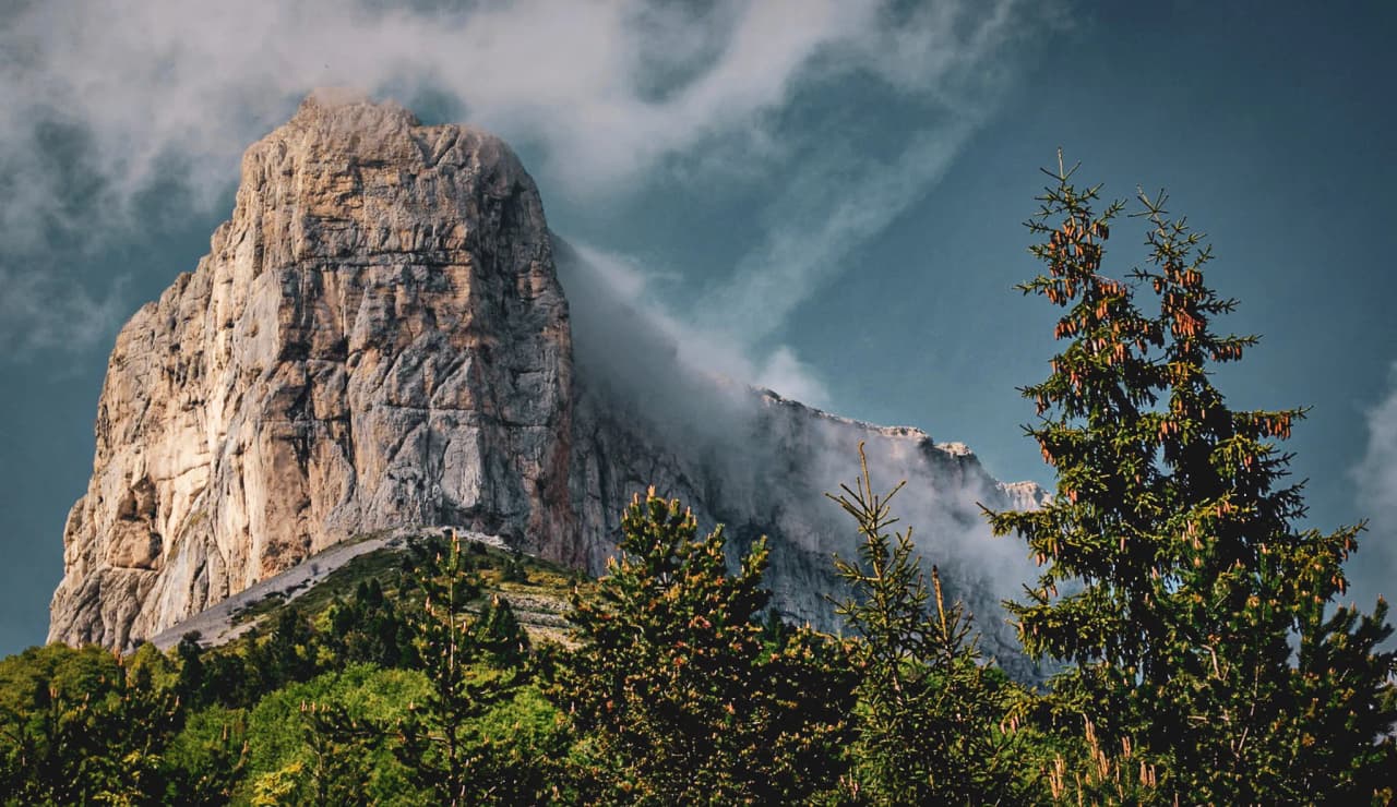 Majestueus uitzicht op de Mont Aiguille, omringd door pijnbomen, onder een blauwe lucht met wolken.