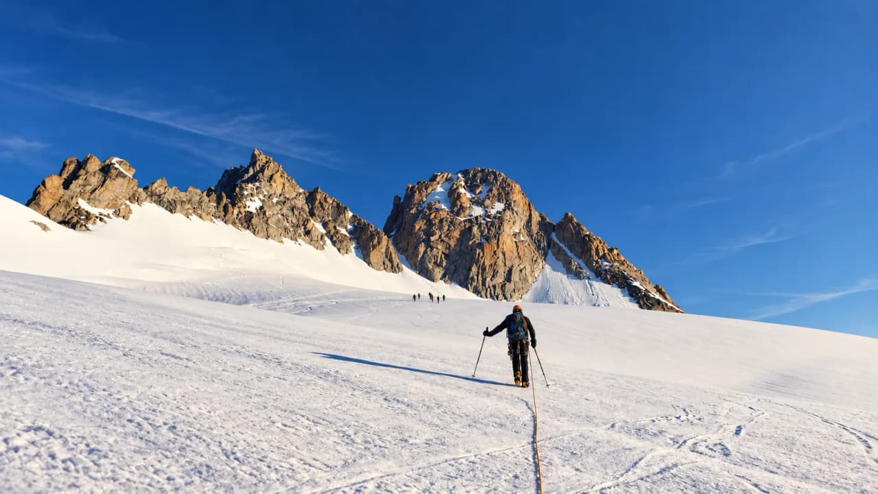 Bergbeklimmer in touwgroep op weg over een gletsjer naar granieten pieken, met een andere touwgroep voor zich.