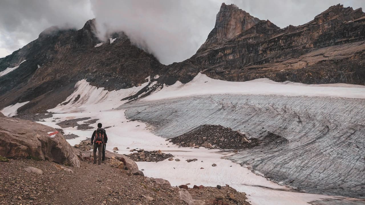 A hiker admires a majestic glacier in the heart of the Alps, under a cloudy sky.