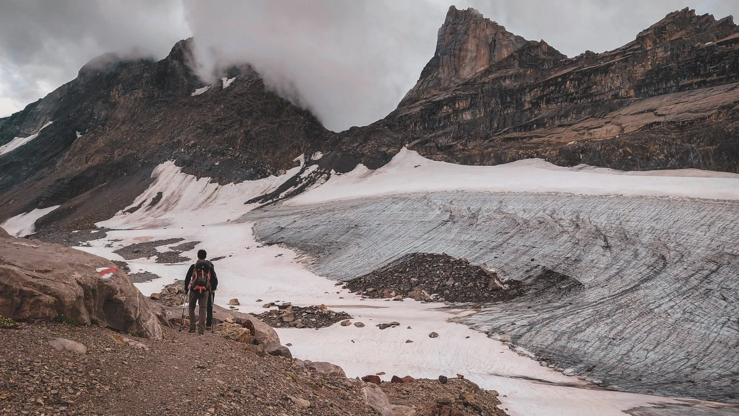 Un randonneur admire un glacier majestueux au cœur des Alpes, sous un ciel nuageux.