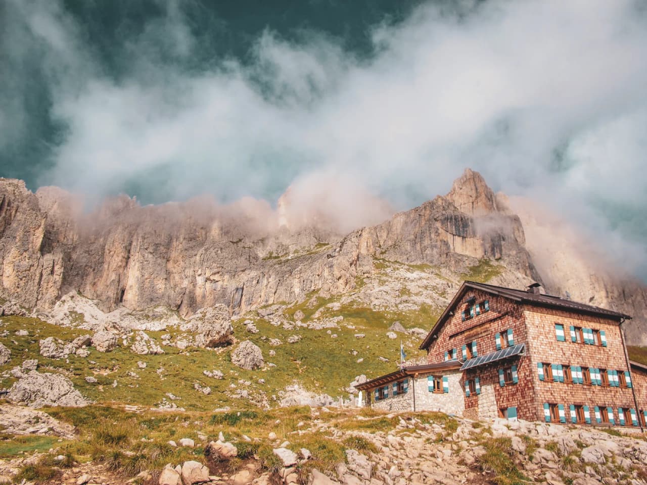 A stone and wood chalet sits in the foreground, backed by majestic cliffs covered in clouds. The building has large windows, allowing you to enjoy the panoramic view of the surrounding countryside.