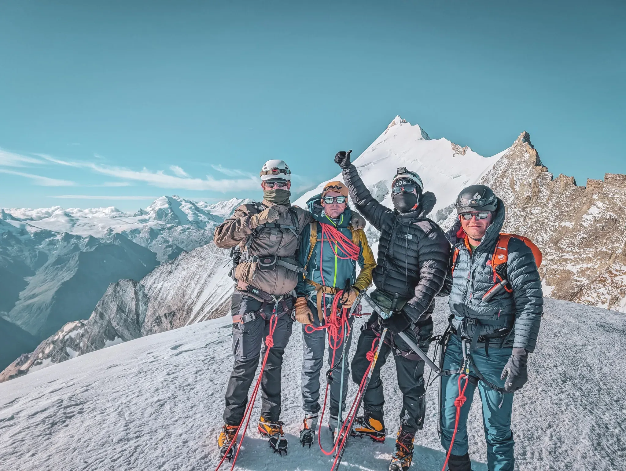 Four climbers on a snow-covered summit, smiling, with breathtaking mountain scenery in the background.