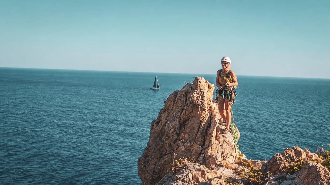 Climbing on a rock overlooking the Mediterranean, with a clear blue sky and a sailing boat in the distance.