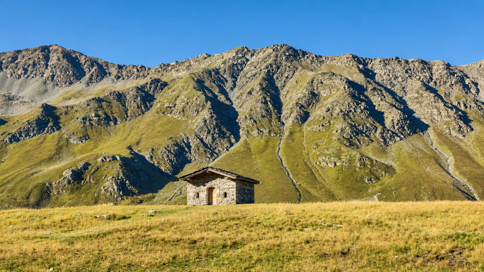 A stone mountain hut stands amidst the lush green mountains, beneath a brilliant blue sky.