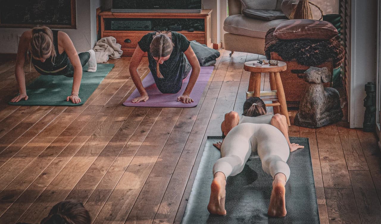 Een groep mensen beoefent yoga in een lichte ruimte met een houten vloer. Drie vrouwen doen verschillende houdingen op hun yogamatjes, één in plankpositie, een ander op haar roze matje en de derde helemaal naar achteren.