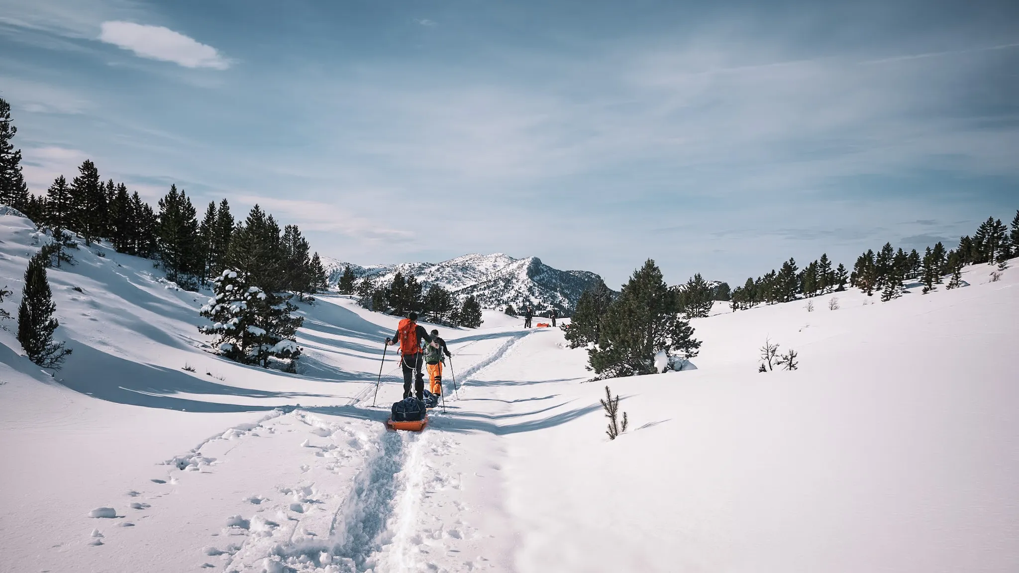 Piste de raquettes en neige, randonneurs avec pulkas sous un ciel bleu, paysage montagneux.