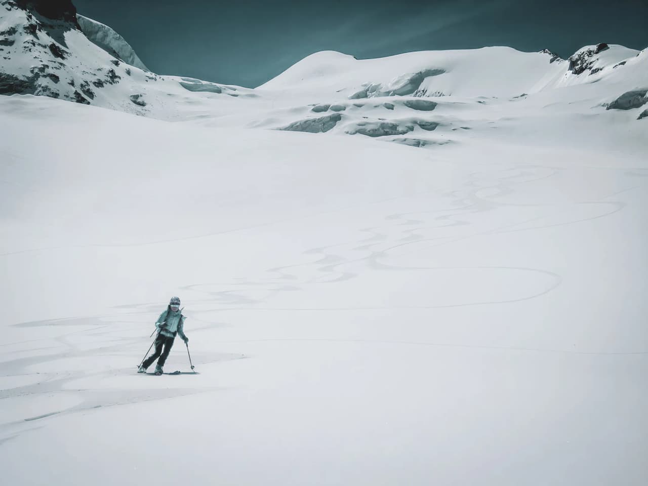skieur solitaire sur un vaste glacier blanc, devant des sommets majestueux sous le ciel bleu.
