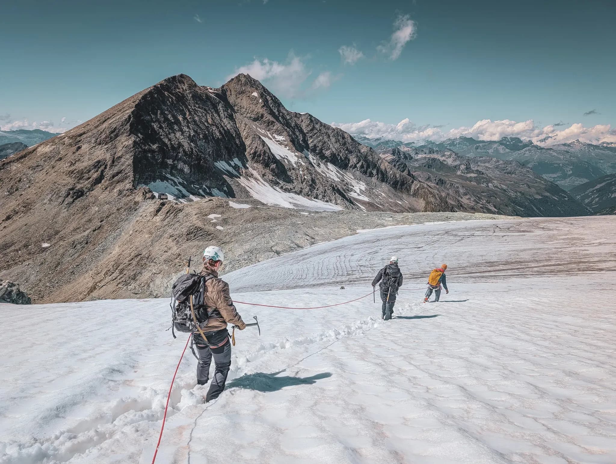 Three climbers advance on a glacier, with majestic peaks in the background.