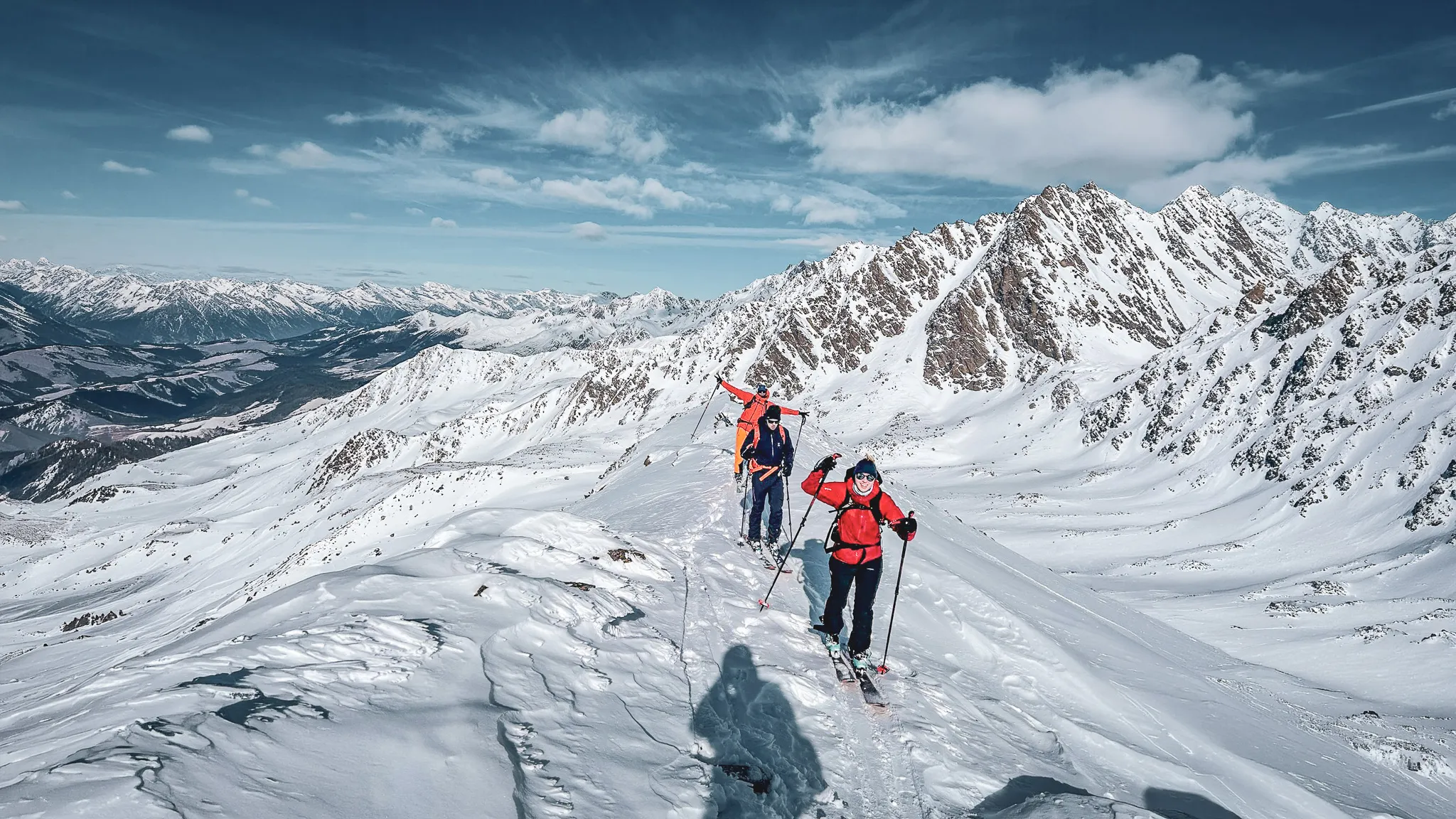 Groupe de skieurs en randonnée alpine sur un sommet enneigé, avec des montagnes majestueuses en arrière-plan.