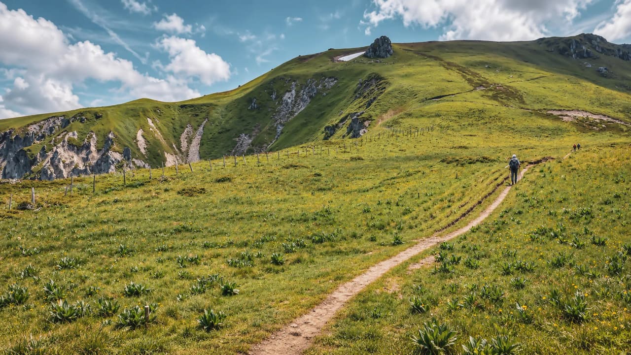Un sentier sinueux traverse un vaste paysage verdoyant sous un ciel bleu parsemé de nuages.