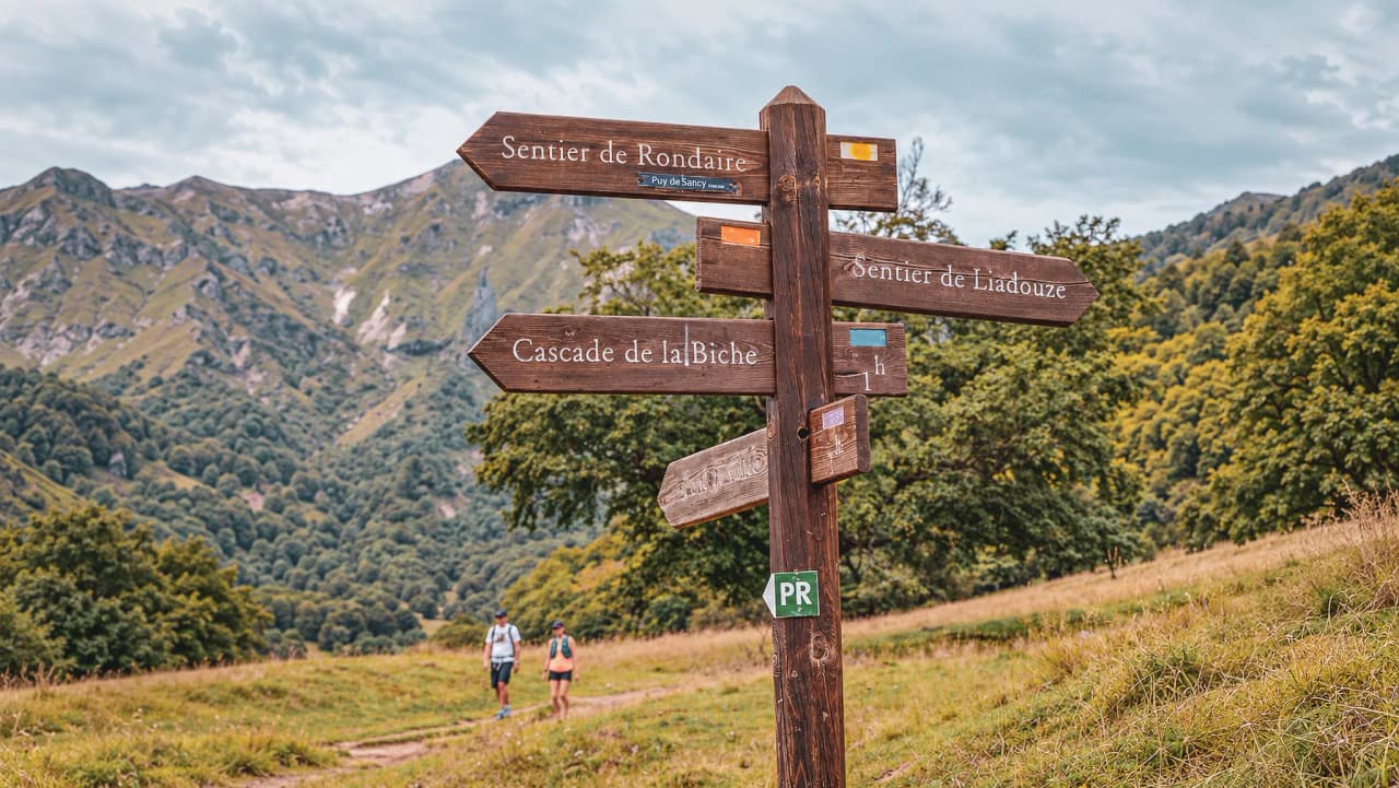 Panneaux indicateurs en bois dans le Massif Central, avec montagnes verdoyantes en arrière-plan.