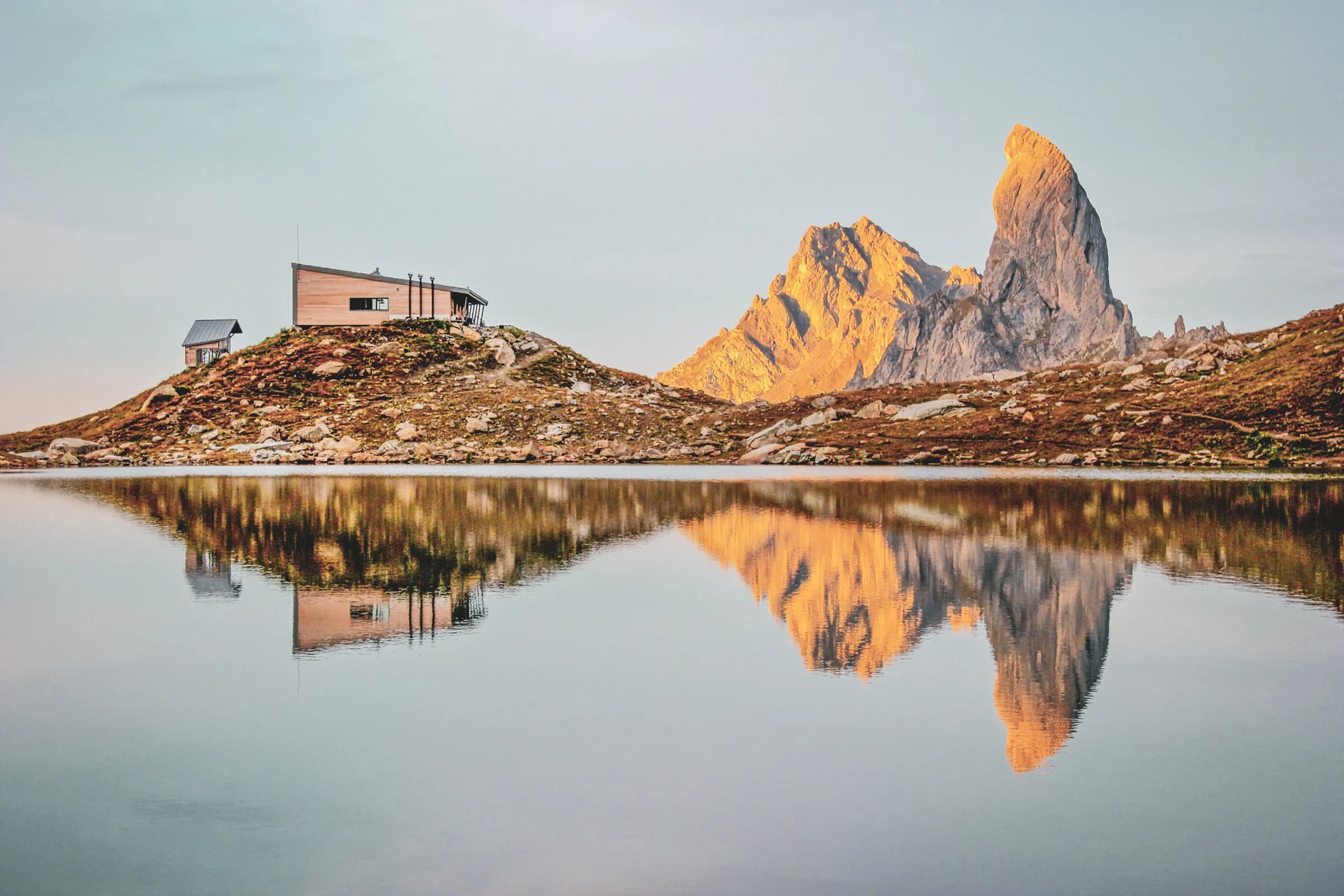 Berghut aan de rand van een meer, met weerspiegeling van de gouden bergtoppen bij zonsopgang. Ontsnapping gegarandeerd.
