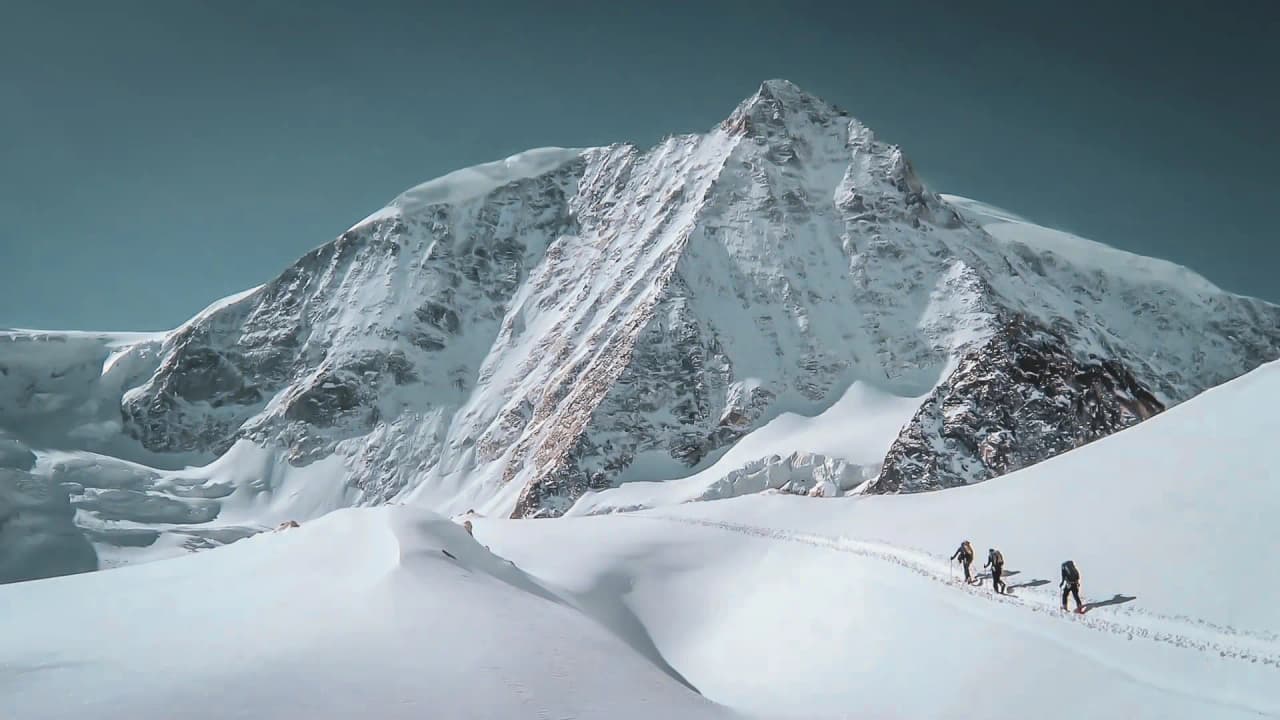 Een indrukwekkend berglandschap, gedomineerd door een ruige besneeuwde top omringd door grote hellingen bedekt met schitterende sneeuw. Aan de voet van de berg zijn een aantal avonturiers uitgerust met sneeuwscooteruitrusting en een lawinepieper onderweg.