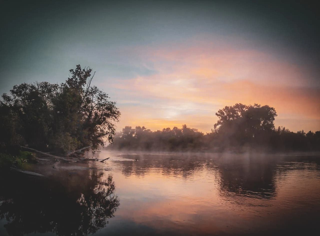 Een landschap aan het water, met een kalm meer dat de zachte kleuren van de zonsopgang weerspiegelt. Weelderige bomen omlijsten de scène, terwijl een lichte nevel boven het water opstijgt en een sfeer van rust creëert.