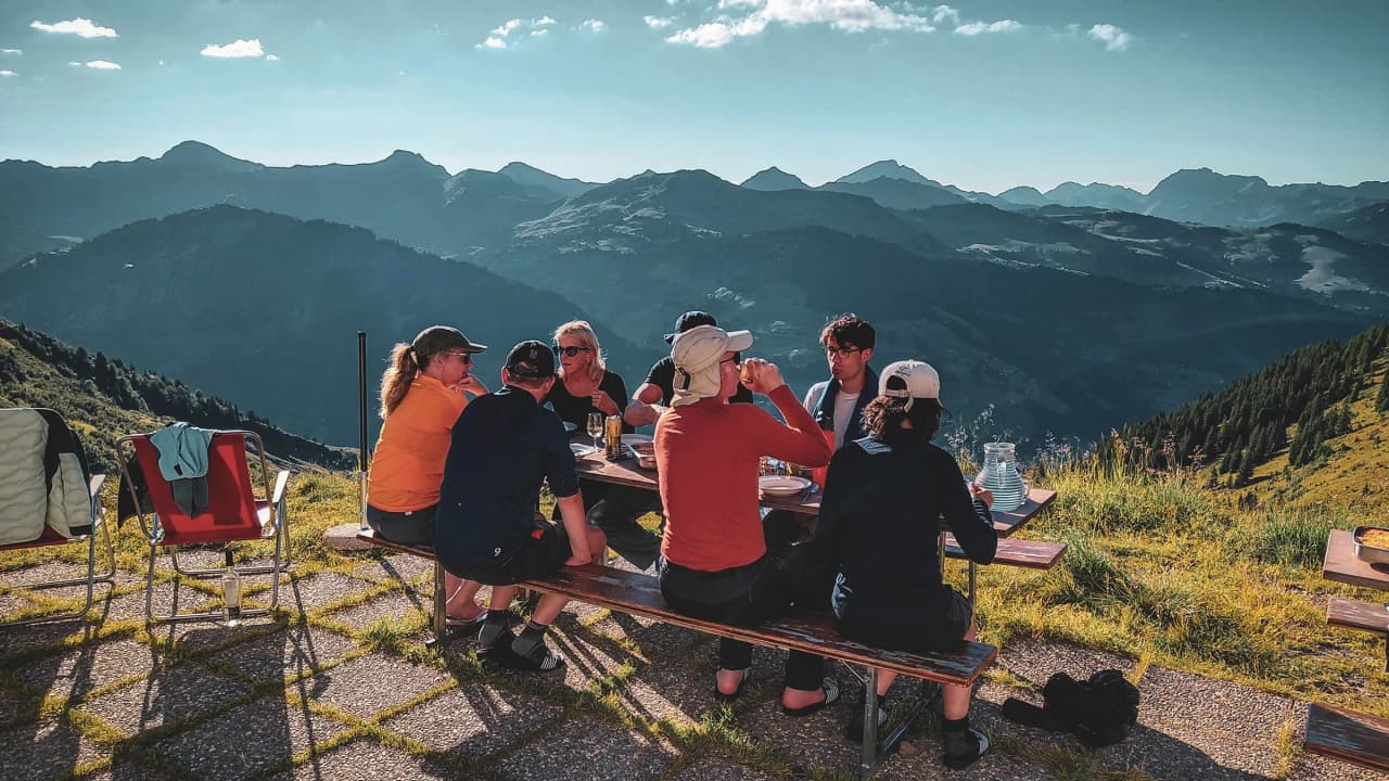 A group gathered round a table outdoors, admiring the mountains and sharing a convivial meal.