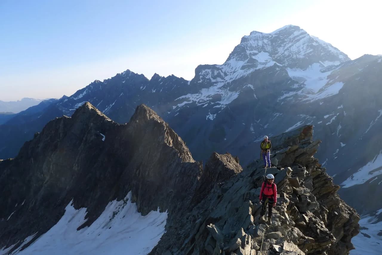 Two climbers are making their way along a rocky ridge facing Mont Blanc, under a bright sky.