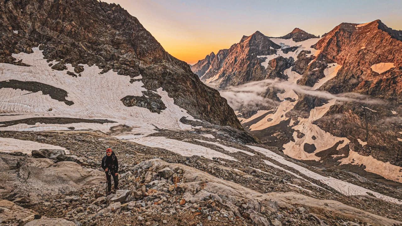 Un alpiniste grimpe sur un sentier rocailleux, entouré de majestueux sommets des Écrins au lever du soleil.