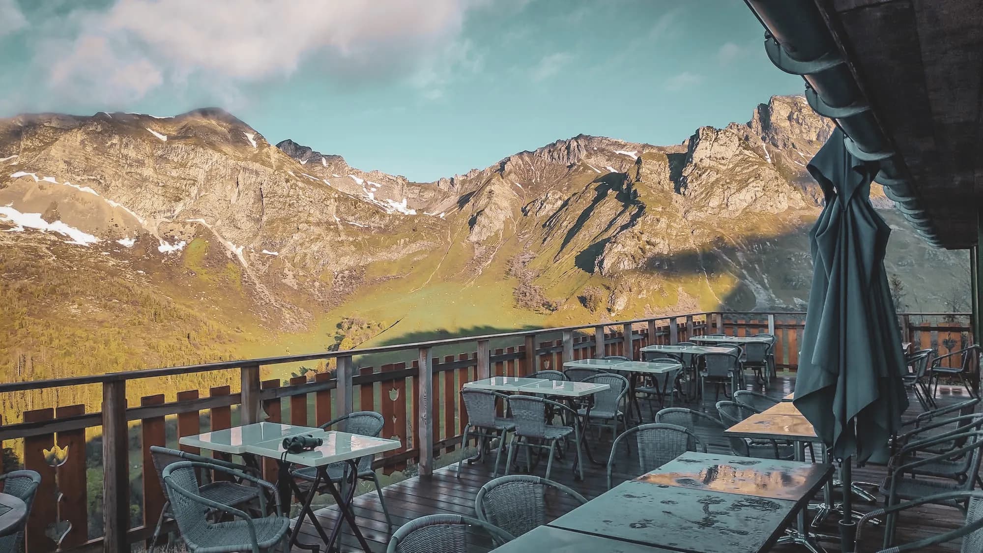 Wooden terrace with tables overlooking majestic mountains, blue skies and clouds.