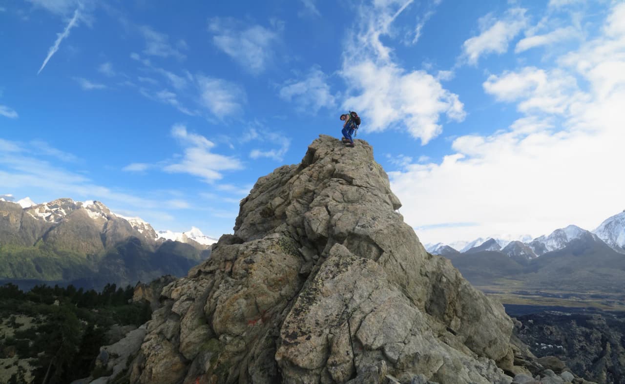 A climber on a majestic crag, facing snow-capped mountains and a brilliant blue sky.