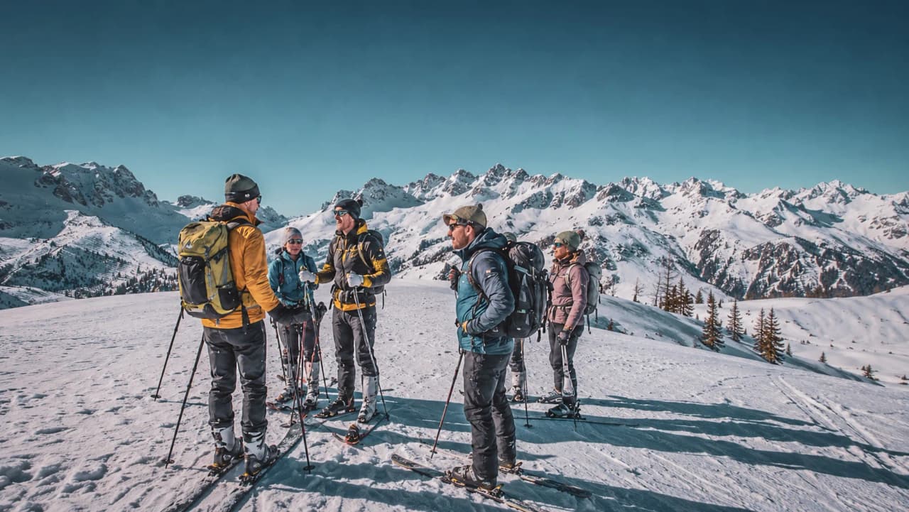 A group of friends on a ski tour, with snow-capped mountains in the background, in an alpine setting.