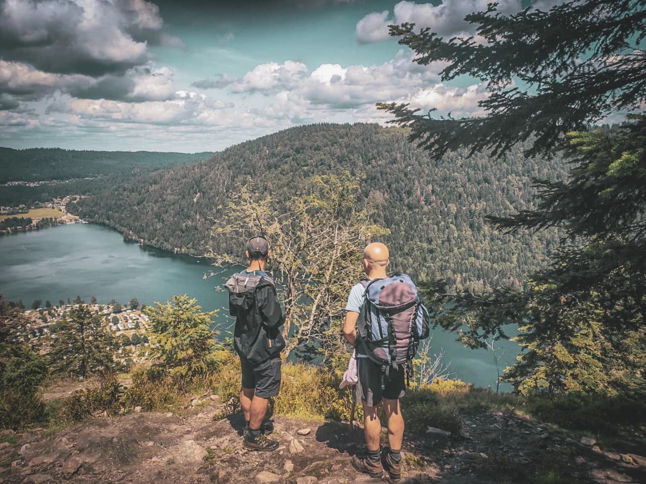 Two hikers admire a green panorama on the edge of a lake in the Vosges under a dramatic sky.