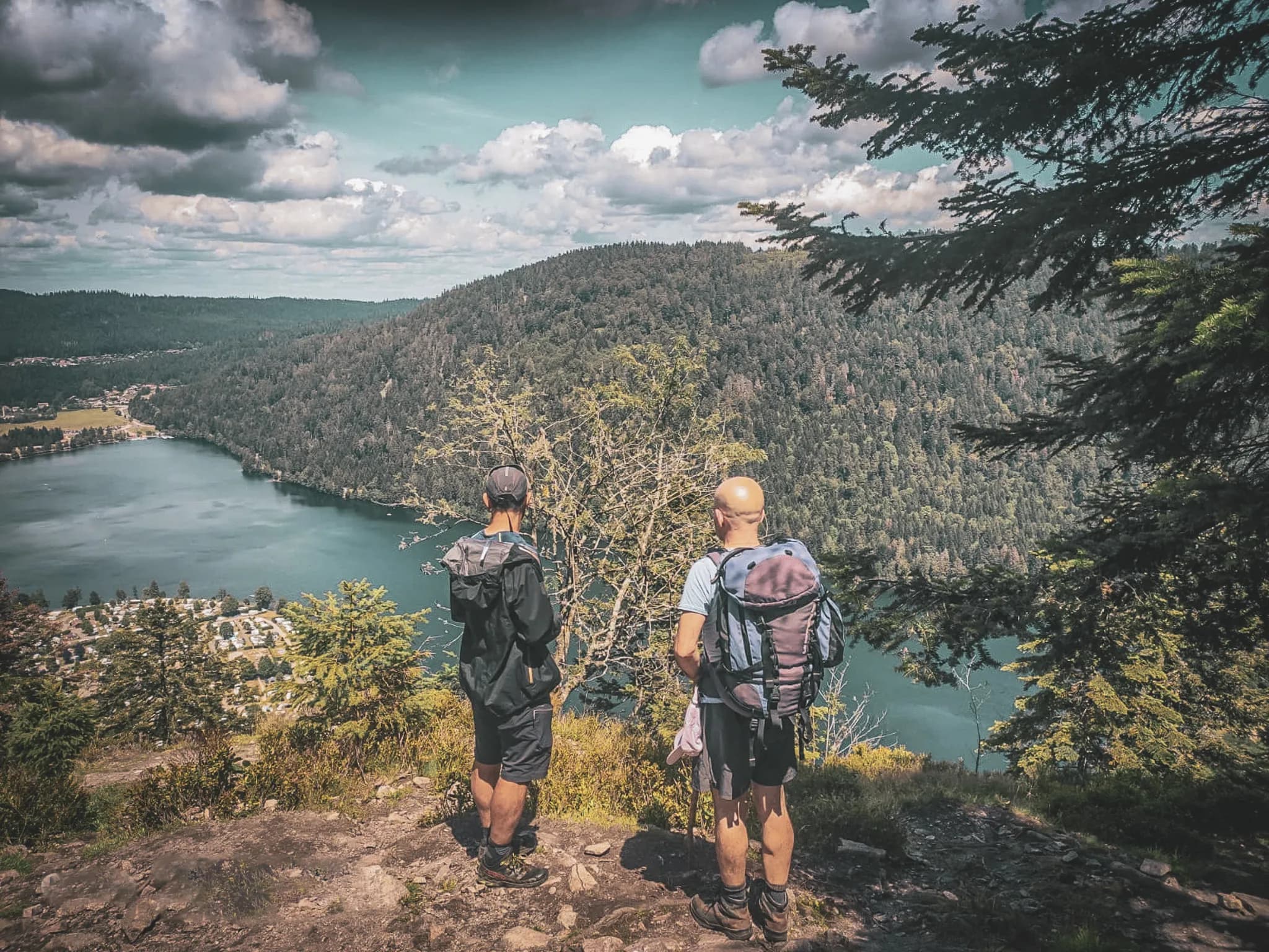 Two hikers admire a green panorama on the edge of a lake in the Vosges under a dramatic sky.