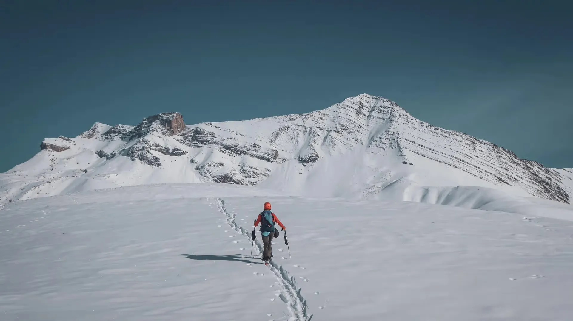 A skier ventures into a snow-covered Alpine landscape, under a brilliant blue sky.