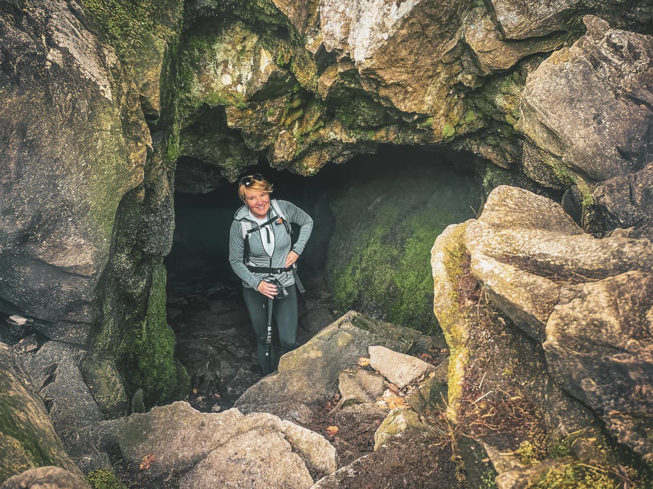 Smiling woman at the entrance to a cave, surrounded by rocks and greenery, inviting adventure.