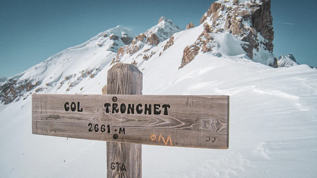 Panneau en bois indiquant le col Tronchet, avec des montagnes enneigées en arrière-plan.