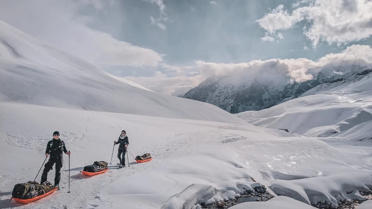 Two snowshoe hikers are pulling pulkas across the snow, against a backdrop of breathtaking mountain scenery.
