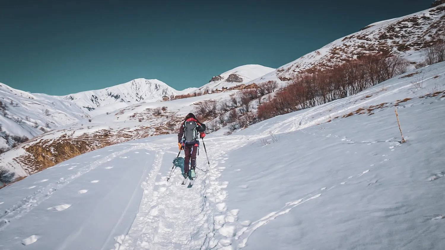 A lone skier makes his way through a snowy mountain landscape under a blue sky.