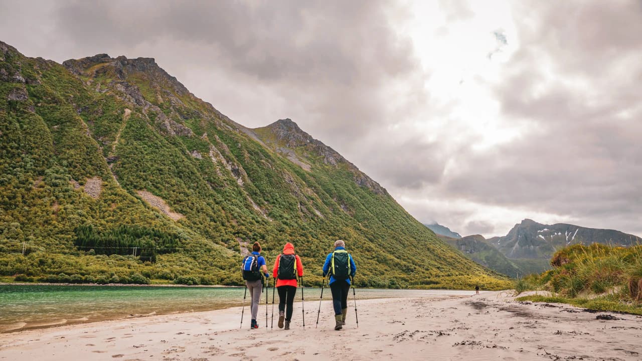 Three hikers walking on a beach, surrounded by green mountains in the Lofoten Islands.