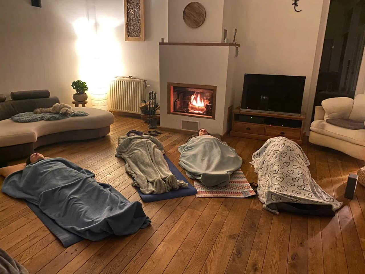 Four participants lie comfortably in a cosy room, next to a fireplace, wrapped in soft blankets, inviting you to relax.