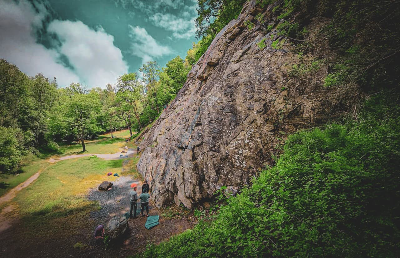 A group of climbers prepare to scale a green rock face under cloudy skies.