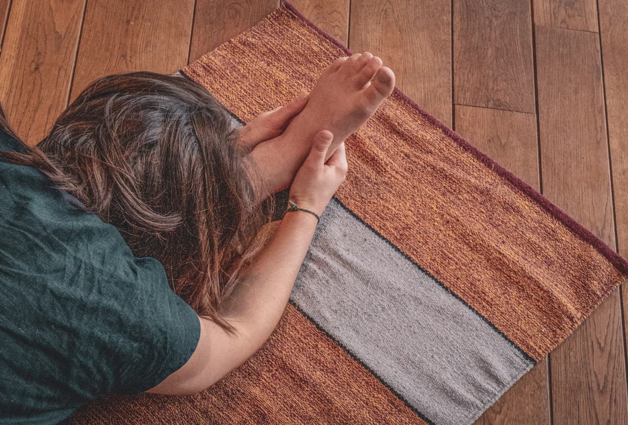 Iemand ligt op een mat en buigt voorover om een voet op te pakken. De greep is zacht en aandachtig, wat duidt op een moment van ontspanning of strekken. De mat bestaat uit verschillende gekleurde banden met verschillende kleurschakeringen.