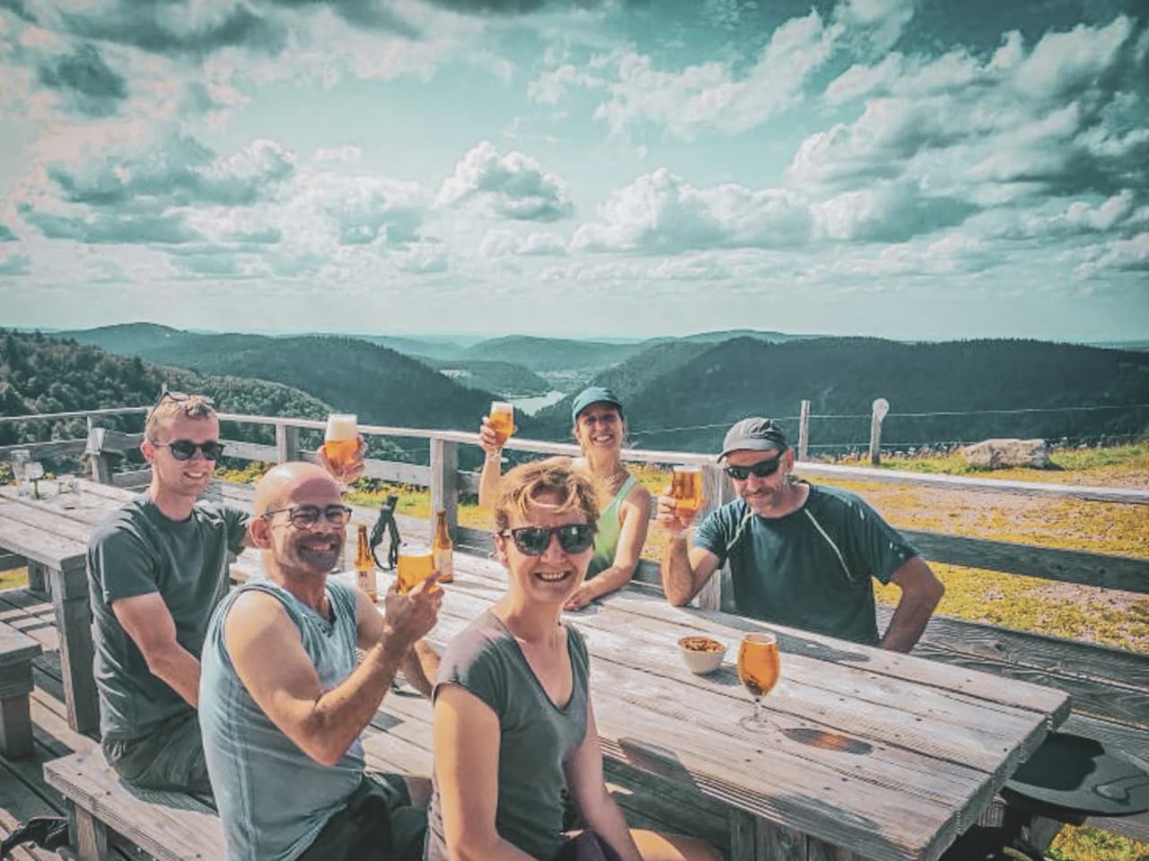A cheerful group on a terrace, raising their glasses in front of a sunny panorama of the Vosges mountains.