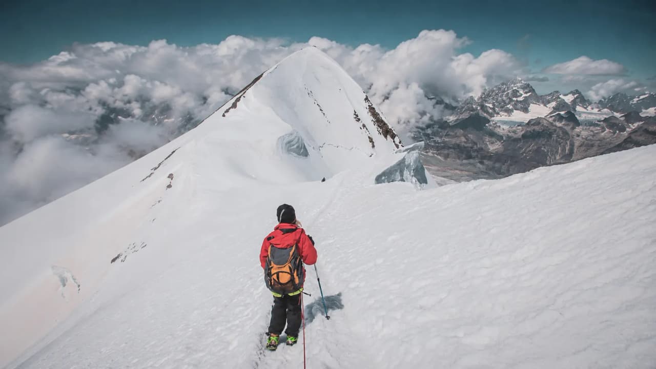 A mountaineer dressed in red on a snow-capped peak, surrounded by majestic clouds and mountains.